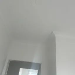 Białe wykończenie narożnika sufitu z listwami dekoracyjnymi, surowy otwór drzwiowy w dolnej części kadru, widok z dołu.