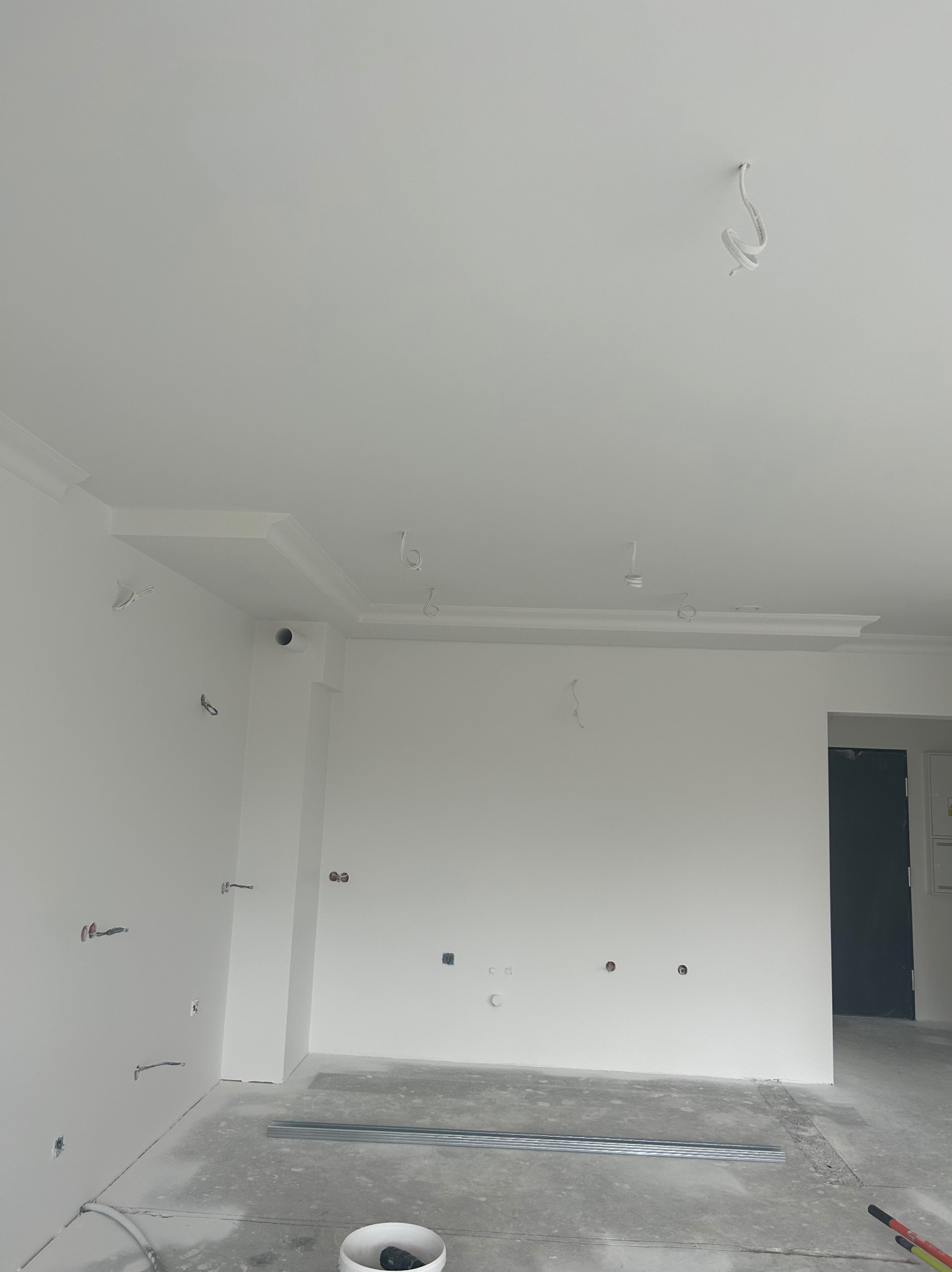 Wnętrze w trakcie wykończenia: białe ściany, listwy dekoracyjne pod sufitem, wystające kable elektryczne, surowa podłoga z narzędziami i profilem metalowym.