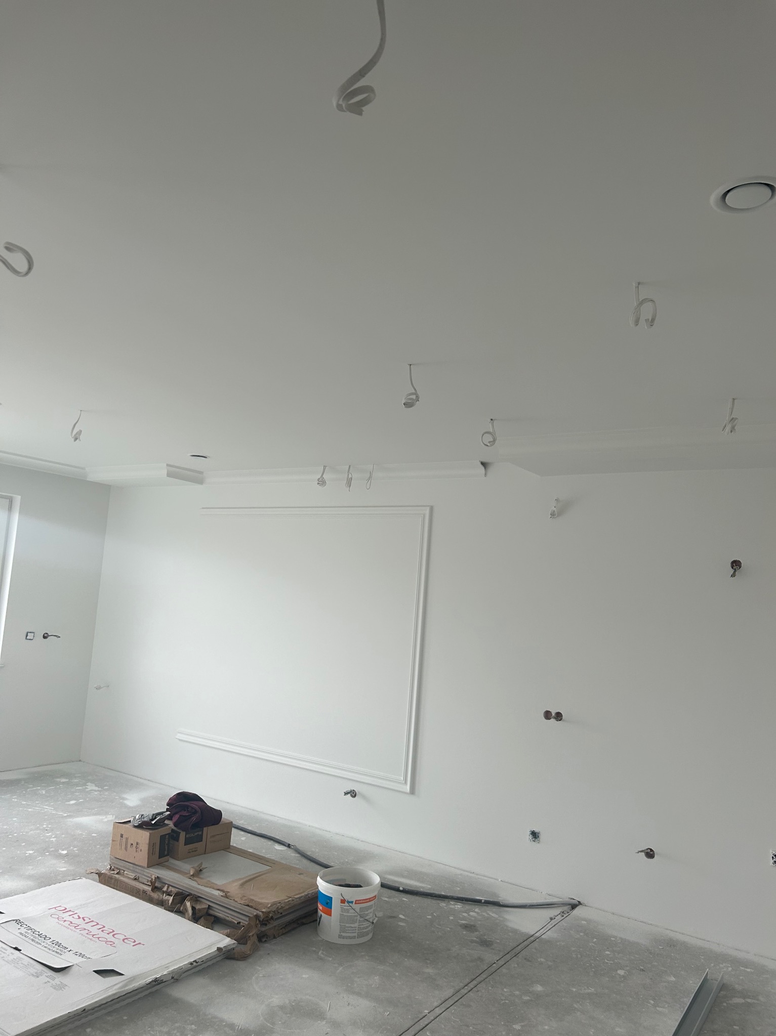 Wnętrze w trakcie prac wykończeniowych: białe ściany z dekoracyjną sztukaterią, przygotowane do montażu oświetlenia, podłoga zabezpieczona folią, widoczne materiały budowlane i narzędzia.