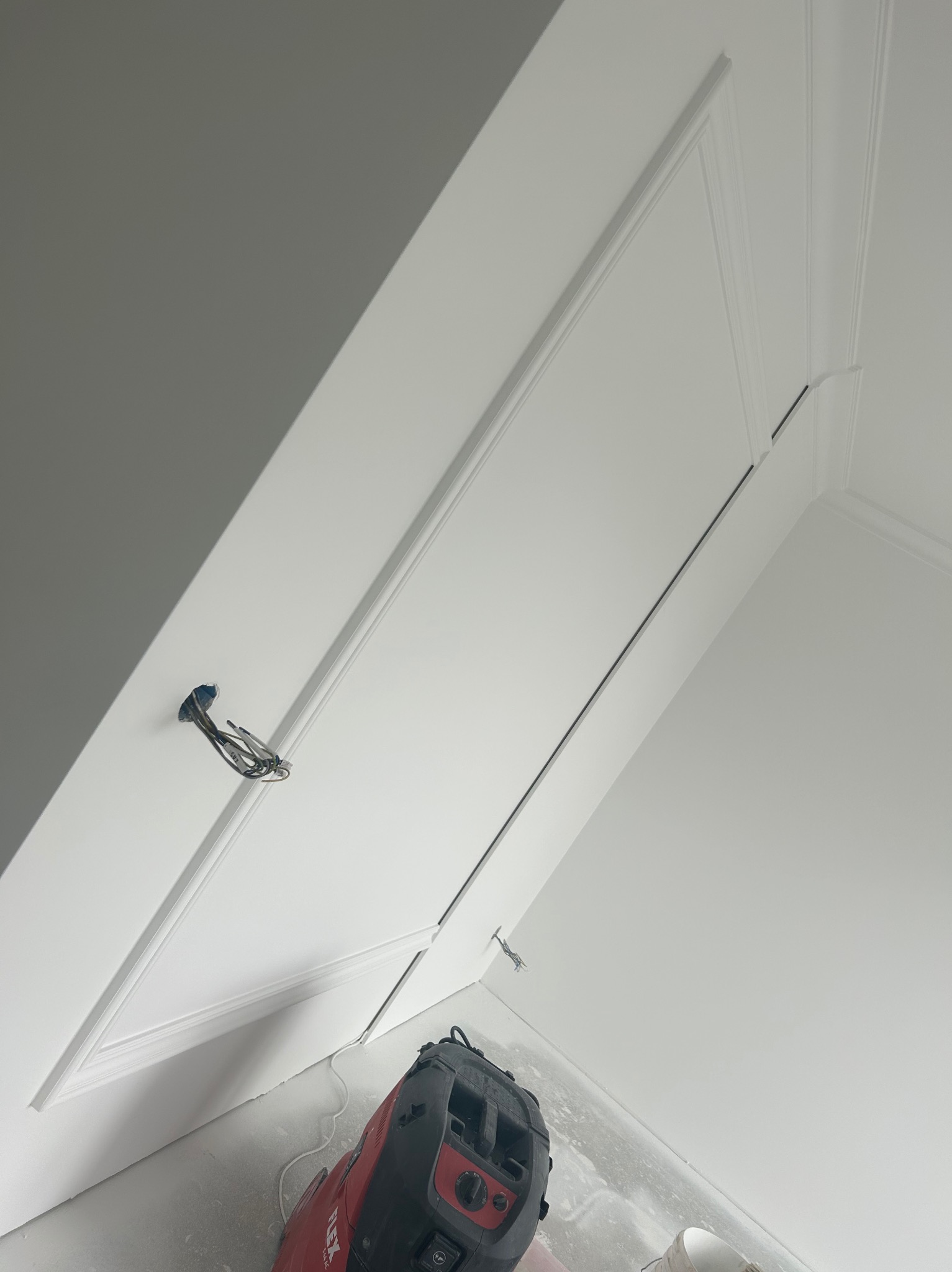 Białe ściany i sufit połączone ozdobnymi listwami, widoczny wystający kabel elektryczny i odkurzacz przemysłowy na podłodze.