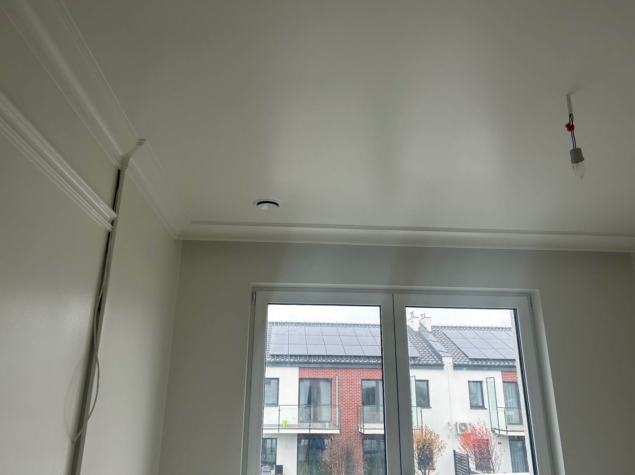 Wnętrze po remoncie: gładka, biała ściana z dekoracyjną listwą przysufitową, okno z widokiem na budynek z panelami słonecznymi na dachu, sufit z zamontowanym oświetleniem punktowym i wiszącą...