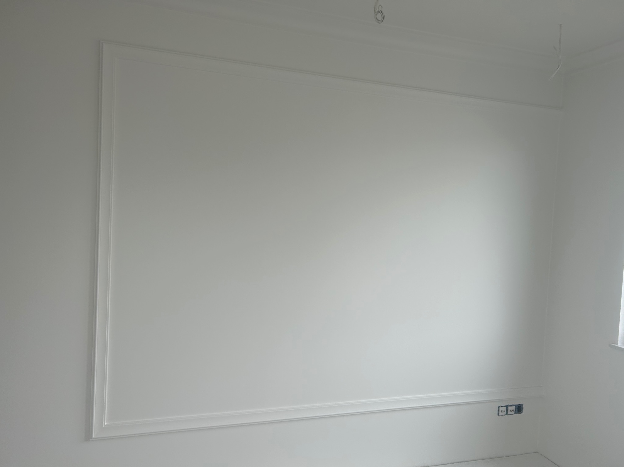 Biała ściana z dekoracyjną ramą i listwami przypodłogowymi, widoczne gniazdka elektryczne, surowy stan wykończenia.