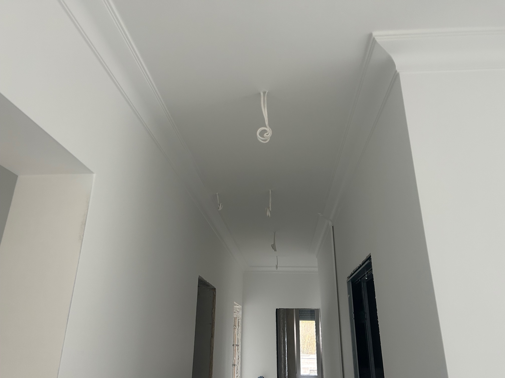 Białe, gładkie ściany i sufit z dekoracyjną listwą przysufitową, widoczne przewody elektryczne zwisające z sufitu, przygotowane do montażu oświetlenia w korytarzu podczas wykańczania wnętrza.