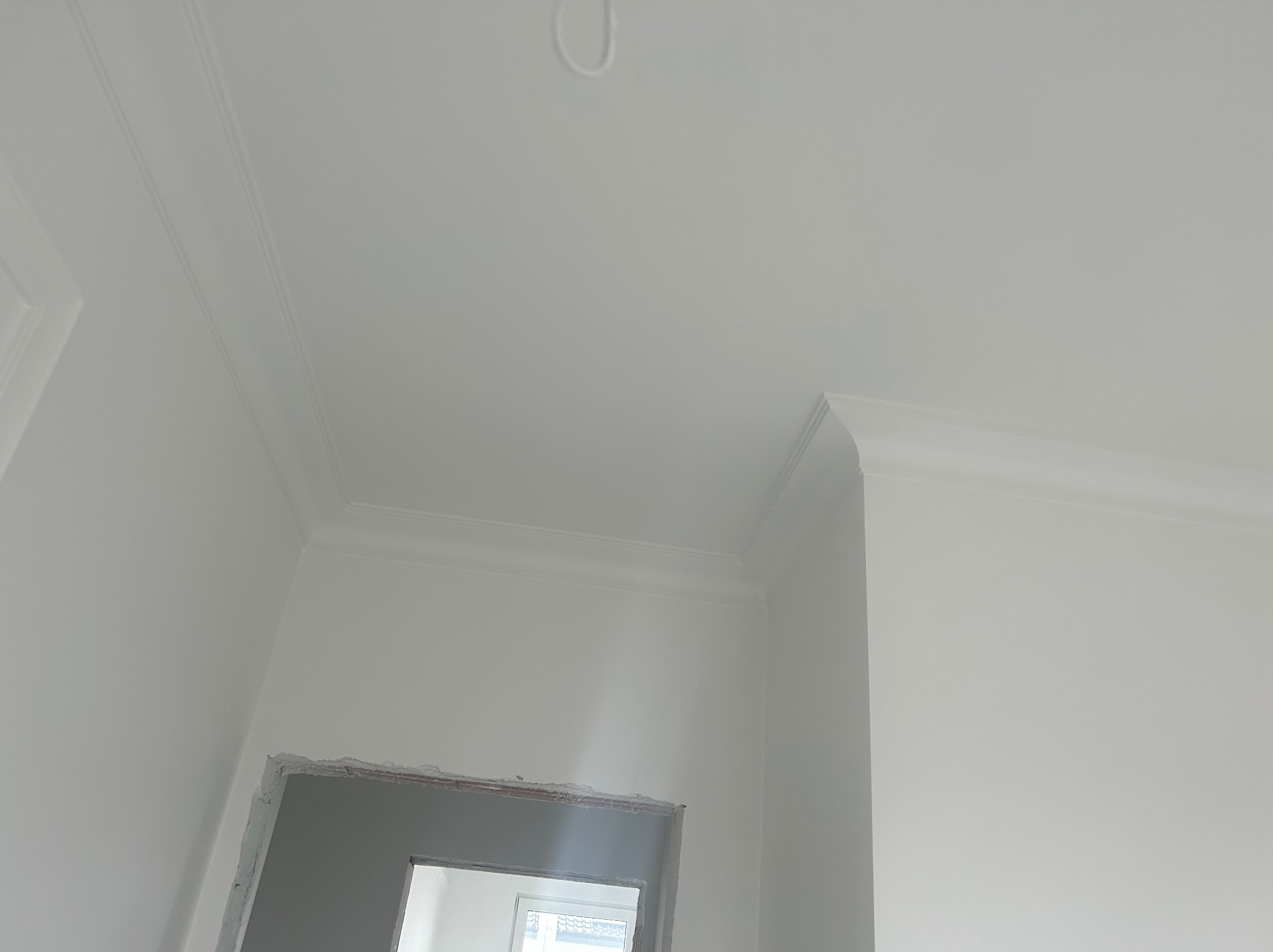 Białe wykończenie narożnika sufitu z listwami dekoracyjnymi, surowy otwór drzwiowy w dolnej części kadru, widok z dołu.