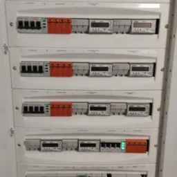 Rozdzielnica elektryczna z zabezpieczeniami przepięciowymi i licznikami energii, widoczne szczegóły podłączeń i oznaczenia producentów.