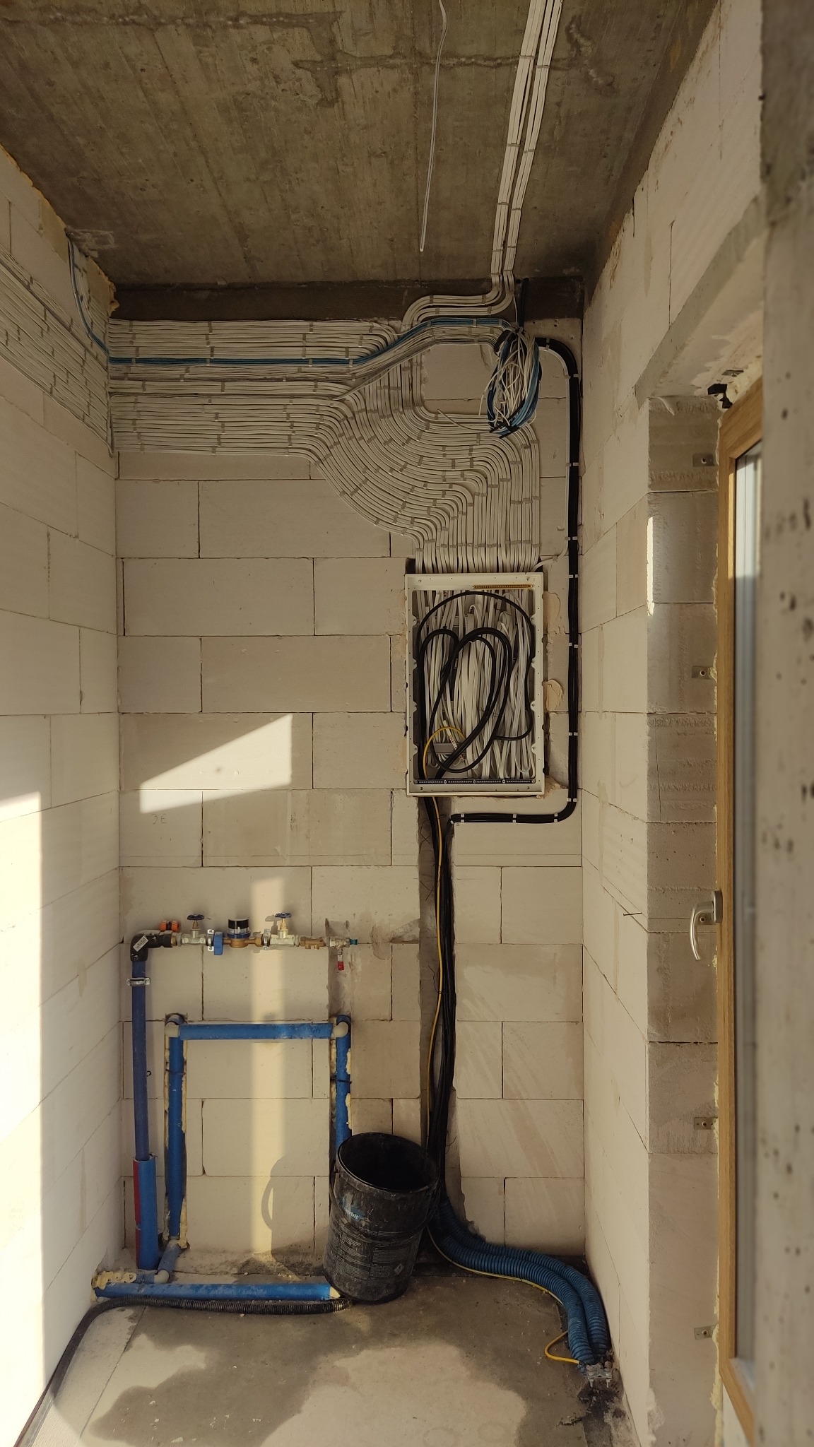 Surowy stan: ściana z betonu komórkowego z licznymi białymi kablami elektrycznymi ułożonymi w równoległe rzędy pod sufitem, skrzynka elektryczna z czarnymi kablami, niebieskie rury instalacji...