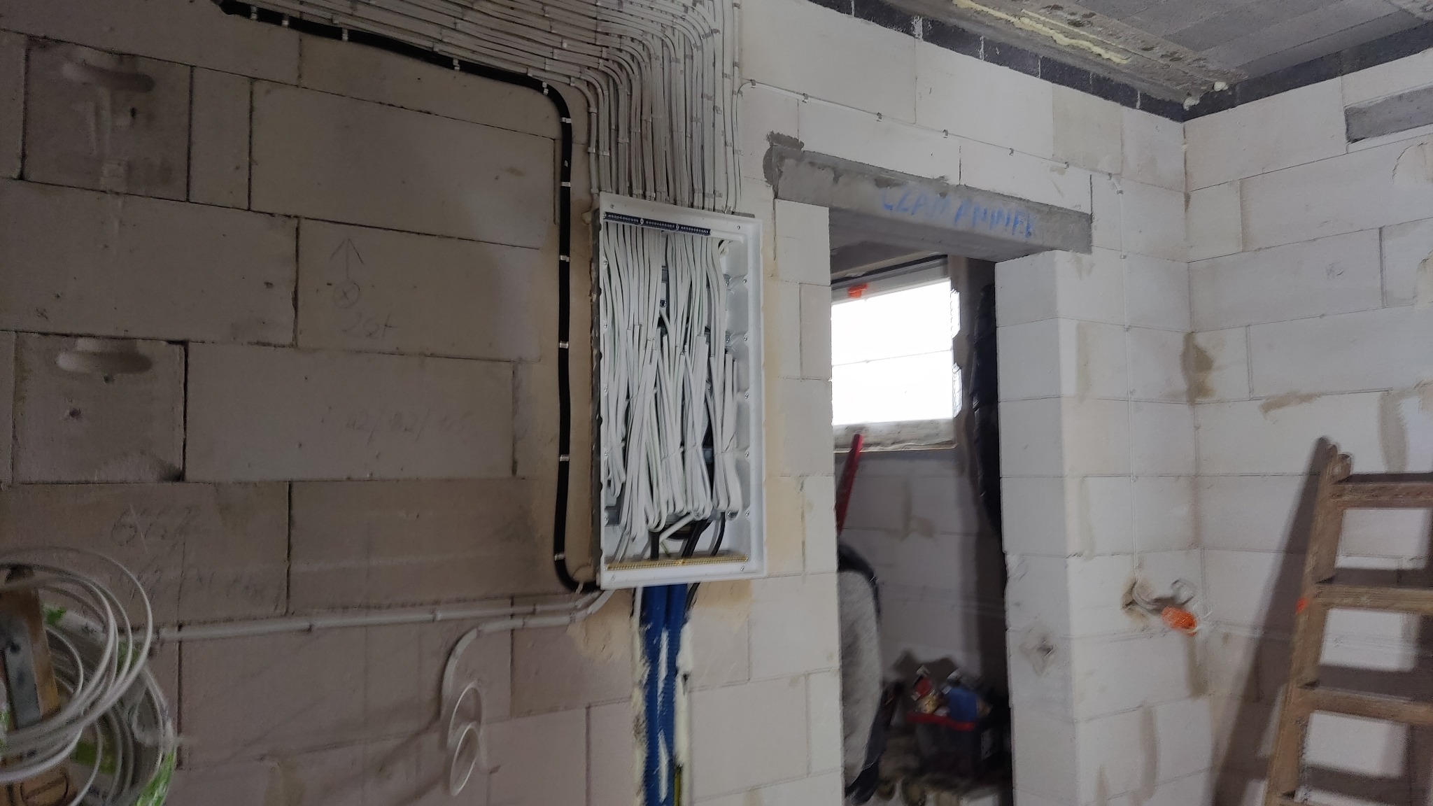 Rozdzielnia elektryczna w trakcie montażu, z licznymi białymi przewodami wychodzącymi z góry i niebieskimi z dołu, umieszczona na ścianie z białych bloczków betonowych w surowym wnętrzu budynku...