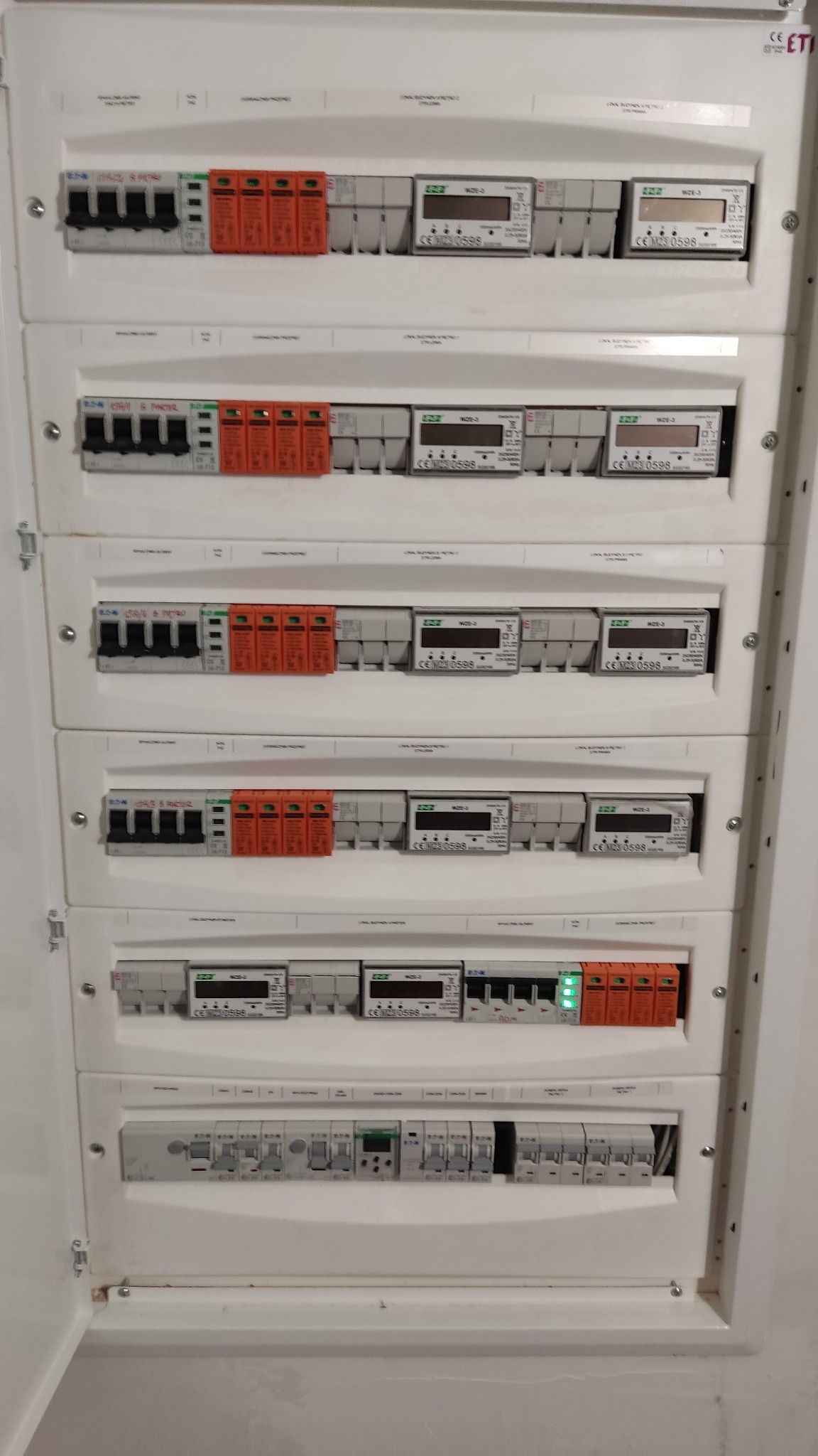 Rozdzielnica elektryczna z zabezpieczeniami przepięciowymi i licznikami energii, widoczne szczegóły podłączeń i oznaczenia producentów.