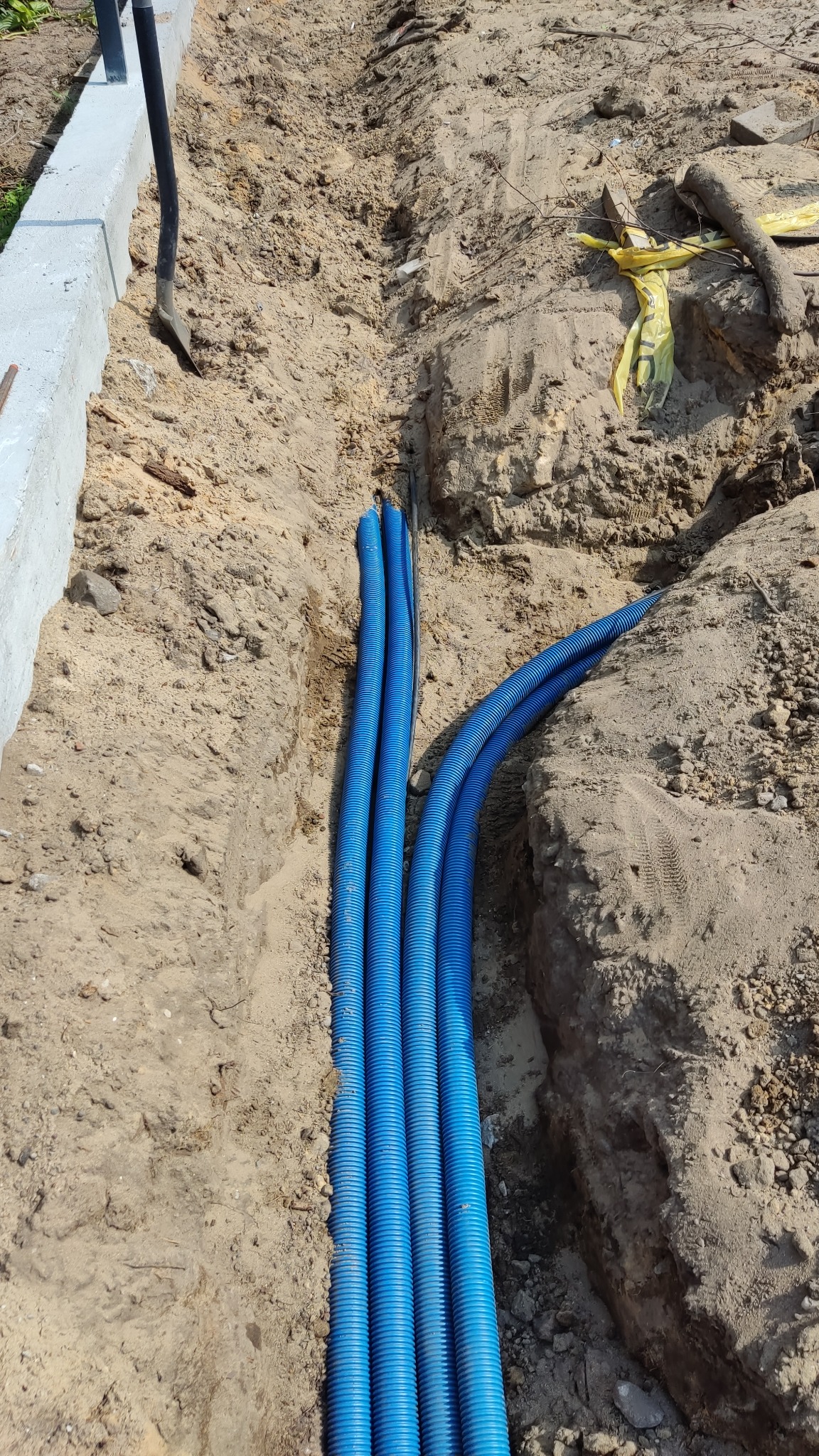 Wykop w ziemi z ułożonymi niebieskimi rurami karbowanymi, prawdopodobnie peszlami, zabezpieczającymi kable; widoczna łopata i fragment betonowego obrzeża.