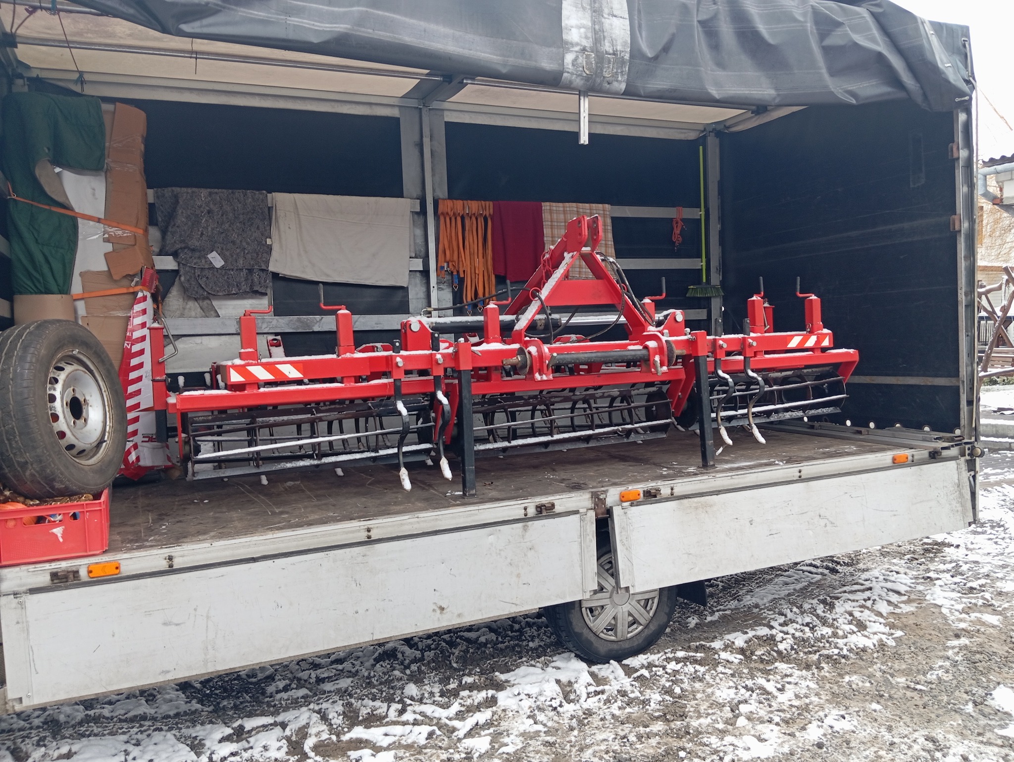 Czerwony agregat uprawowy w skrzyni ładunkowej samochodu dostawczego, zabezpieczony podczas transportu, obok zapasowe koło i inne przedmioty.