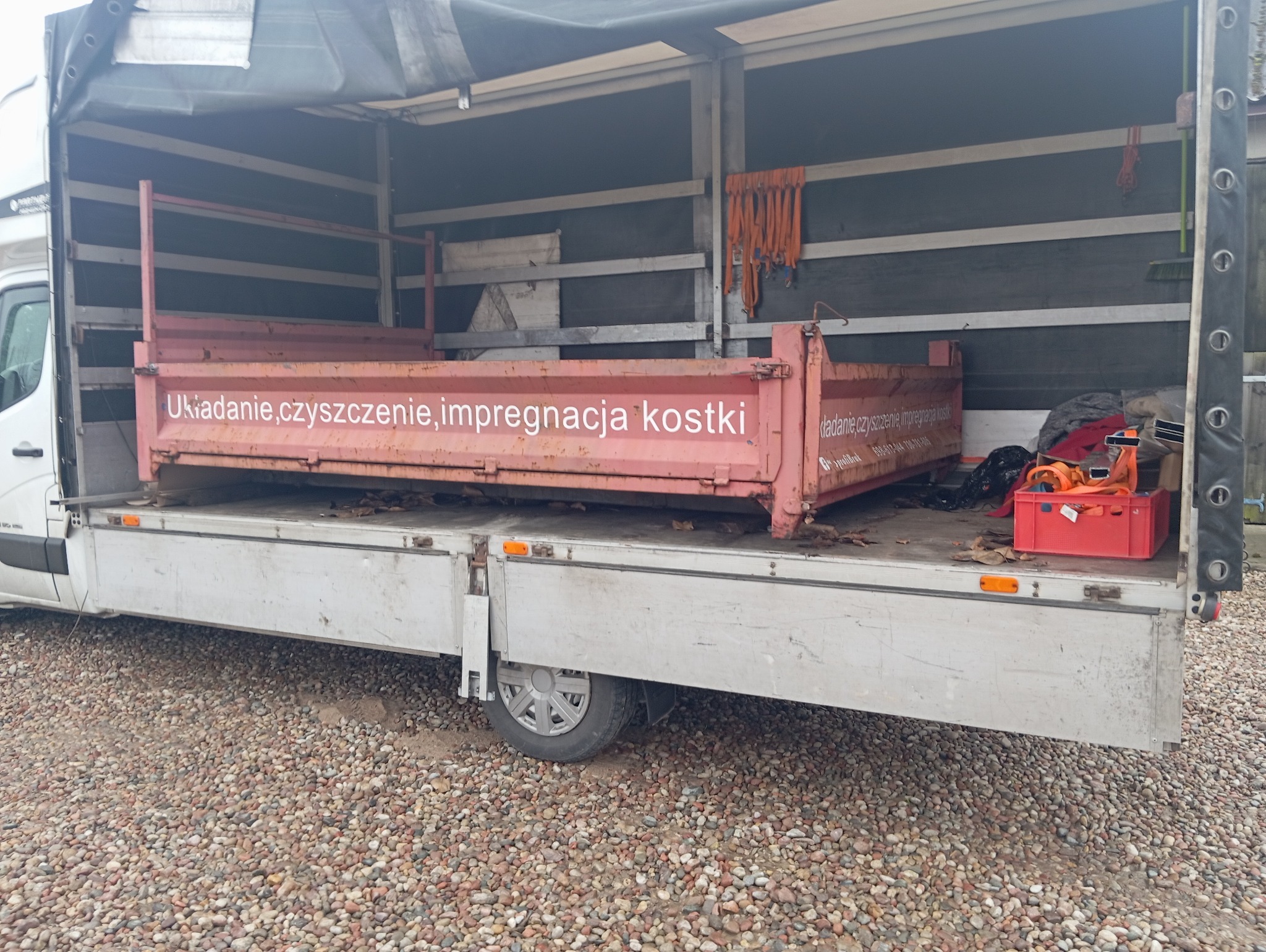 Otwarta skrzynia ładunkowa z napisem 'Układanie, czyszczenie, impregnacja kostki' załadowana na platformę samochodu dostawczego, na tle żwirowego podłoża.