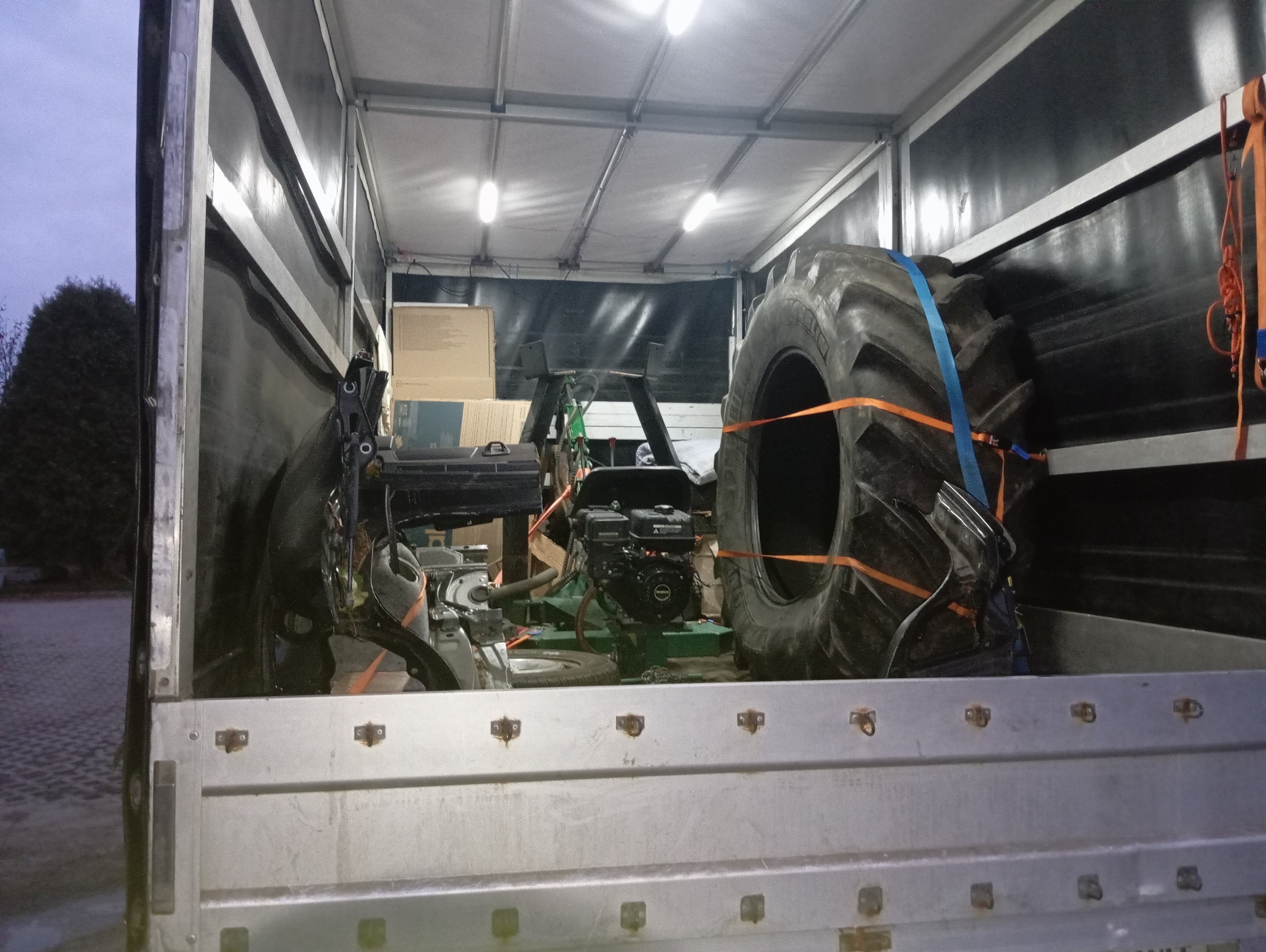 Wnętrze przyczepy z załadowanym silnikiem, oponą rolniczą i elementami konstrukcyjnymi, zabezpieczonymi pasami transportowymi.