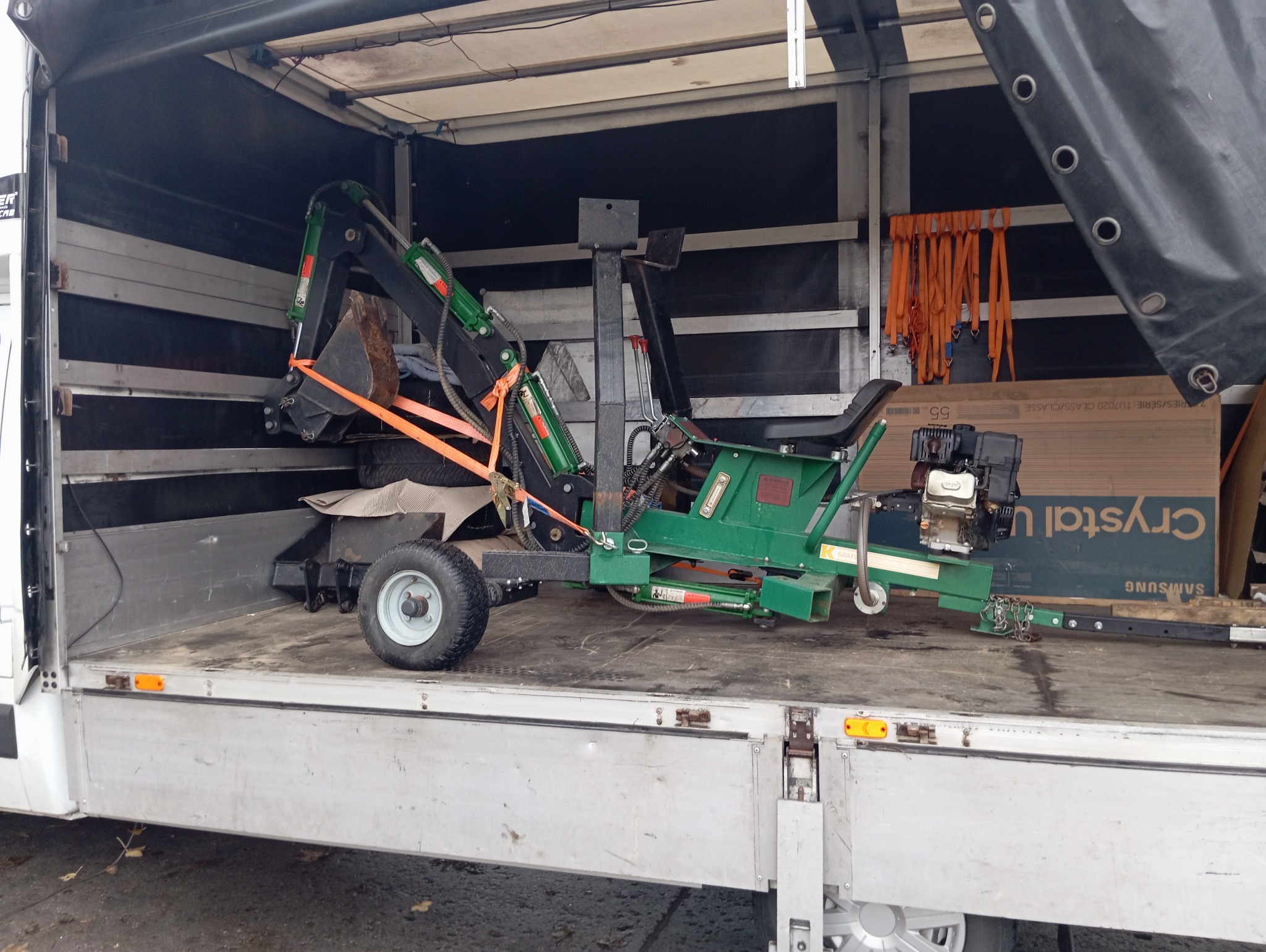 Mini koparka zabezpieczona pomarańczowymi pasami wewnątrz skrzyni ładunkowej białego busa dostawczego, widoczne koło i fragment skrzyni telewizora Samsung.