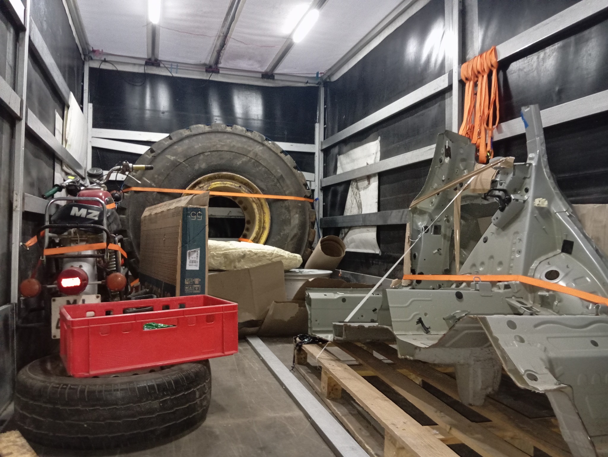 Wnętrze przestrzeni ładunkowej z zabezpieczonym ładunkiem: opona od traktora, motocykl MZ, karoseria samochodu i inne przedmioty, wszystko spięte pomarańczowymi pasami.