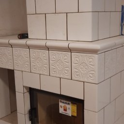 ROBERT OLEKSY - Biały kominek kaflowy z ozdobnymi kaflami, czarną szybą paleniska i naklejką informacyjną producenta Hajduk