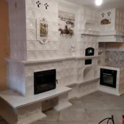 Biały, kaflowy piec kuchenny z kominkiem i piekarnikiem, zdobiony obrazkami, w jasnym pomieszczeniu