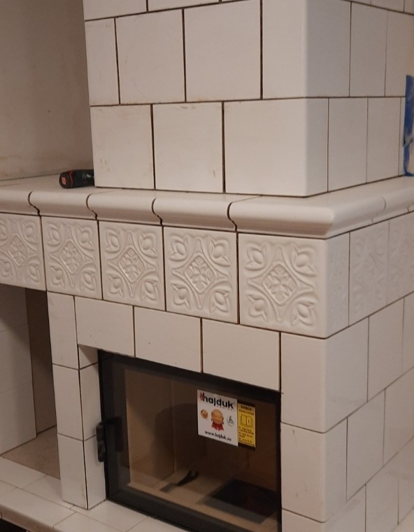 Biały kominek kaflowy z ozdobnymi kaflami, czarną szybą paleniska i naklejką informacyjną producenta Hajduk