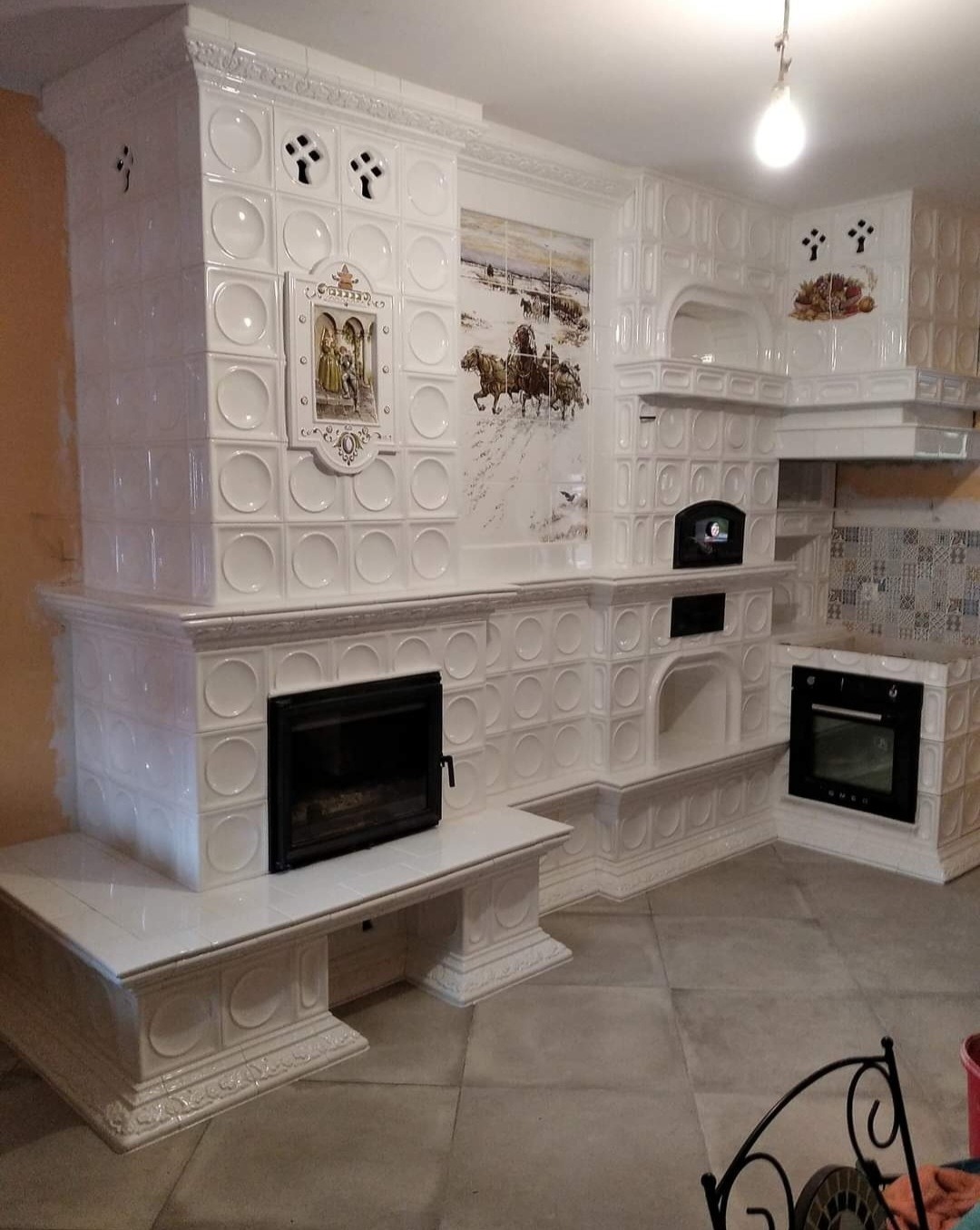 Biały piec kaflowy z kominkiem i zabudowaną kuchenką, zdobiony obrazkami i ornamentami, widok z boku na tle szarej podłogi.