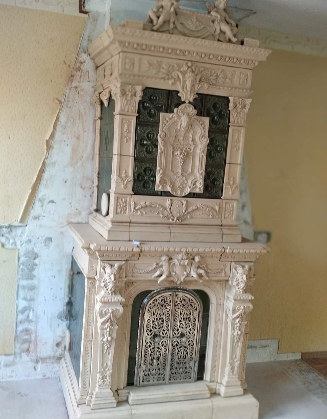 Ozdobny, beżowy piec kaflowy z rzeźbieniami aniołków i roślinnymi motywami, widoczny w trakcie remontu pomieszczenia z odsłoniętą ścianą.