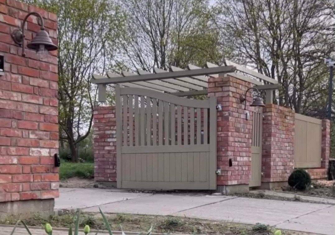 Brama wjazdowa z ceglanymi słupkami i drewnianą pergolą, oświetlona lampą w stylu retro, prowadząca na posesję.