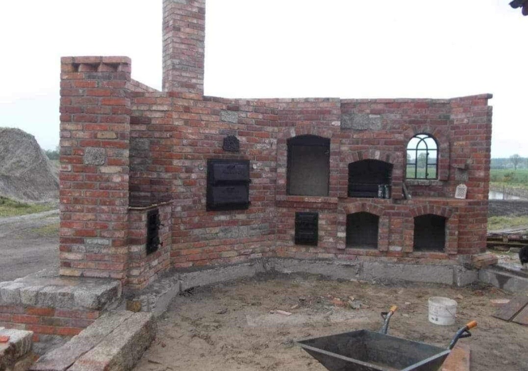 Wnętrze ceglanej konstrukcji z kominem, z otworami na palenisko i piekarnik, w trakcie budowy, widoczna taczka i wiadro na podłożu.