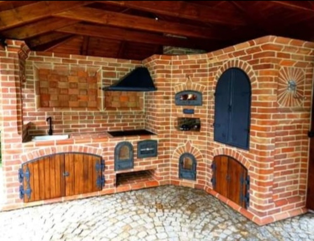 Murowana kuchnia zewnętrzna z cegły klinkierowej pod zadaszeniem w Tuliszkowie, z piecem chlebowym, paleniskiem i zlewem, wykończona drewnianymi drzwiczkami i kamienną posadzką.