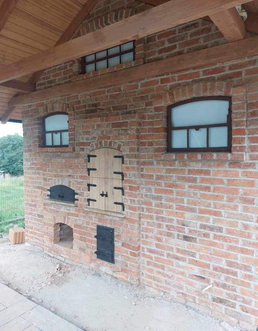 Murowany piec zewnętrzny z cegły z drewnianymi elementami i czarnymi okuciami, widoczne okienka i drzwiczki.