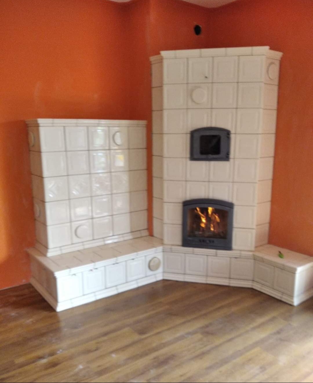 Biały piec kaflowy z paleniskiem, umieszczony w rogu pomarańczowego pomieszczenia, z widocznymi płomieniami wewnątrz.