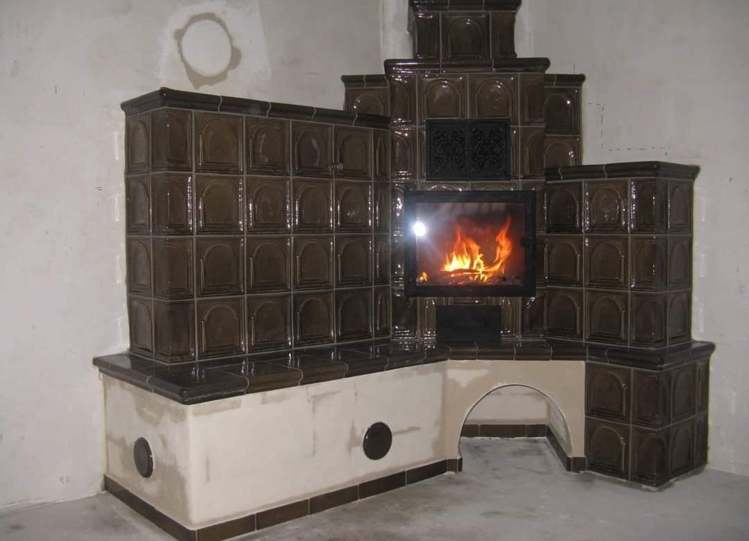 Nowo wybudowany piec kaflowy z brązowymi kaflami, widoczny ogień w palenisku, surowe ściany pomieszczenia.