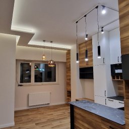 Santi Łukasz Burkiewicz - Nowoczesne oświetlenie sufitowe w mieszkaniu w Bydgoszczy, widoczne drewniane panele podłogowe i zabudowa kuchenna z elementami drewna i czerni.