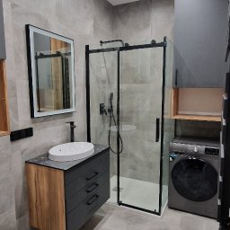 Santi Łukasz Burkiewicz - Nowoczesna łazienka w odcieniach szarości z kabiną prysznicową typu walk-in, umywalką nablatową na szafce z szufladami o wykończeniu drewnopodobnym, pralką i podświetlanym lustrem.