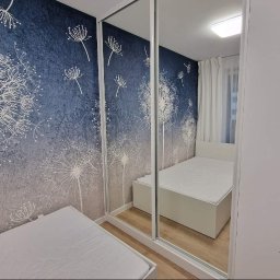 Santi Łukasz Burkiewicz - Sypialnia z łóżkiem i szafą przesuwną z lustrzanymi drzwiami, na ścianie tapeta w dmuchawce w odcieniach szarości i błękitu, widoczne odbicie drugiego łóżka.