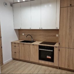 Santi Łukasz Burkiewicz - Nowoczesna, jasna kuchnia z drewnianymi frontami szafek dolnych i białymi górnymi, czarny zlew i płyta indukcyjna wkomponowane w blat, drewnopodobny panel ścienny nad blatem.