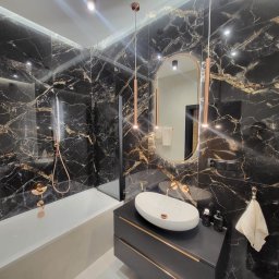 Santi Łukasz Burkiewicz - Elegancka łazienka z czarnymi płytkami imitującymi marmur, armaturą w kolorze różowego złota i owalnym lustrem.
