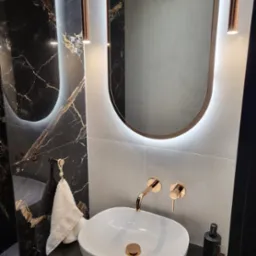 Nowoczesna łazienka z umywalką nablatową, złotą armaturą i podświetlanym lustrem na tle czarnych i szarych płytek imitujących marmur.