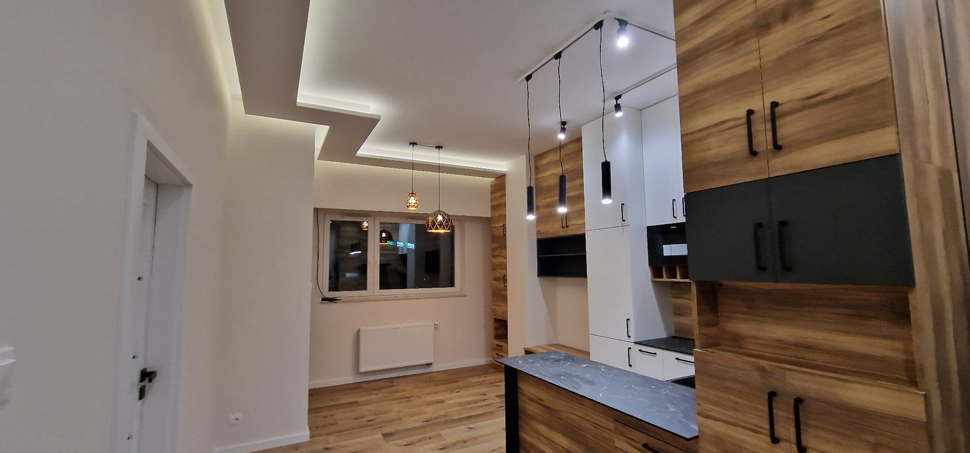 Nowoczesne oświetlenie sufitowe w mieszkaniu w Bydgoszczy, widoczne drewniane panele podłogowe i zabudowa kuchenna z elementami drewna i czerni.