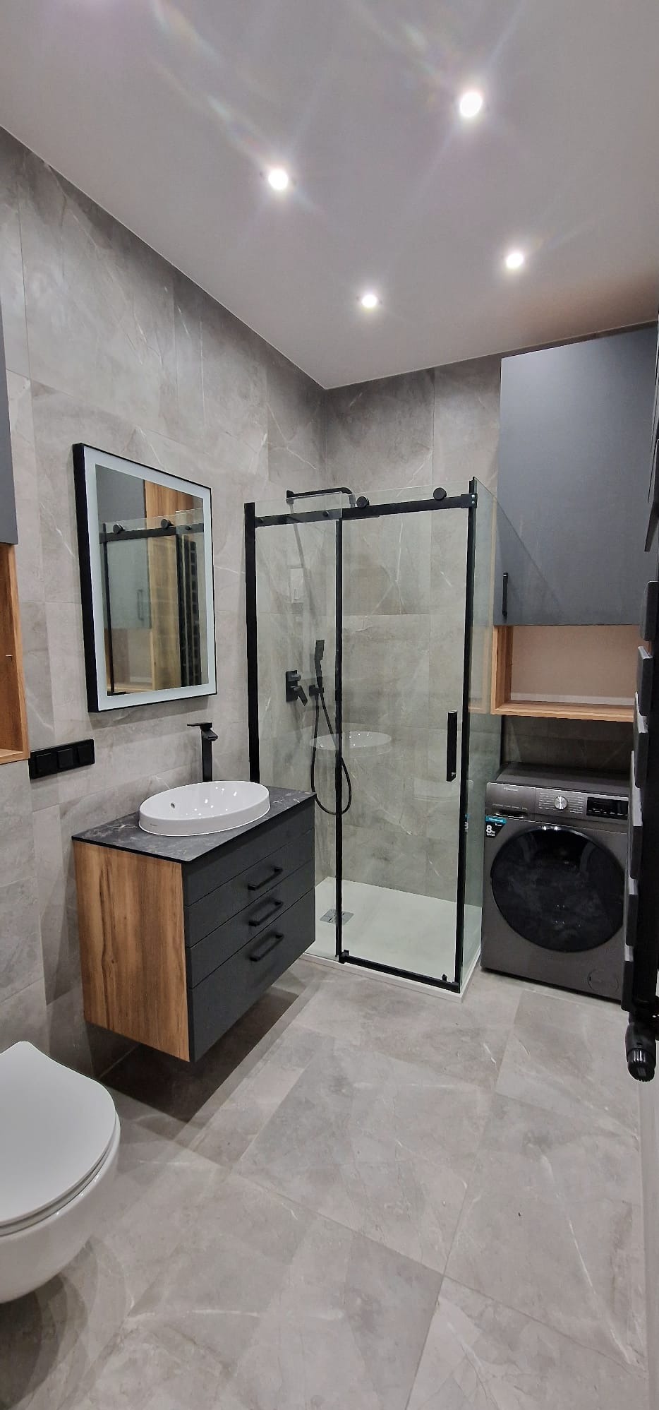 Nowoczesna łazienka w odcieniach szarości z kabiną prysznicową typu walk-in, umywalką nablatową na szafce z szufladami o wykończeniu drewnopodobnym, pralką i podświetlanym lustrem.