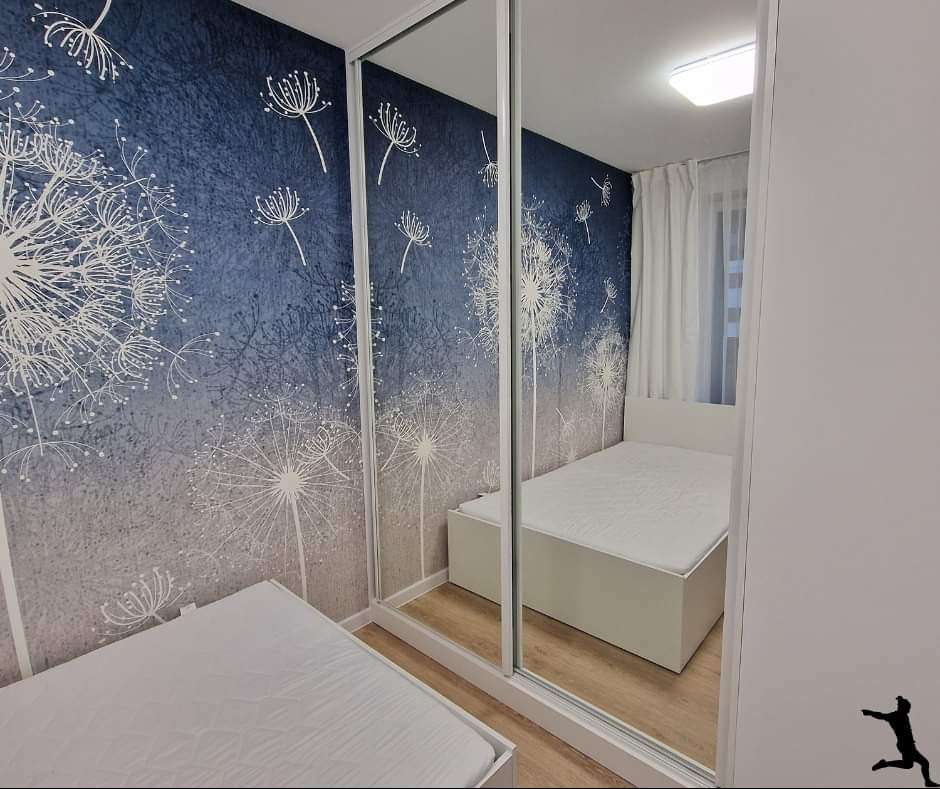 Sypialnia z łóżkiem i szafą przesuwną z lustrzanymi drzwiami, na ścianie tapeta w dmuchawce w odcieniach szarości i błękitu, widoczne odbicie drugiego łóżka.