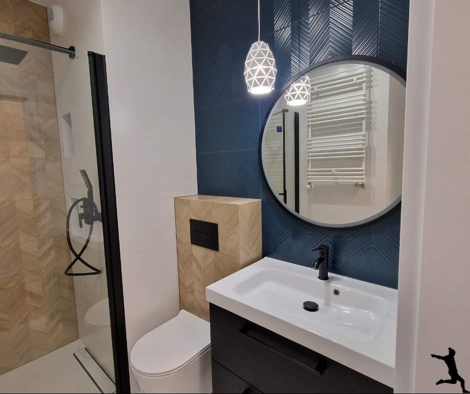 Nowoczesna łazienka z kabiną prysznicową typu walk-in, podwieszaną toaletą z obudową imitującą drewno, umywalką nablatową na czarnej szafce i okrągłym lustrem z czarną ramą, oświetlona wiszącą...