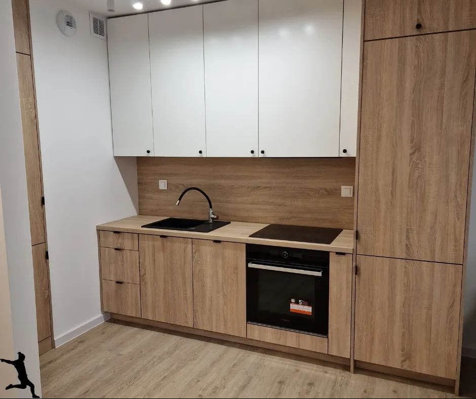 Nowoczesna, jasna kuchnia z drewnianymi frontami szafek dolnych i białymi górnymi, czarny zlew i płyta indukcyjna wkomponowane w blat, drewnopodobny panel ścienny nad blatem.