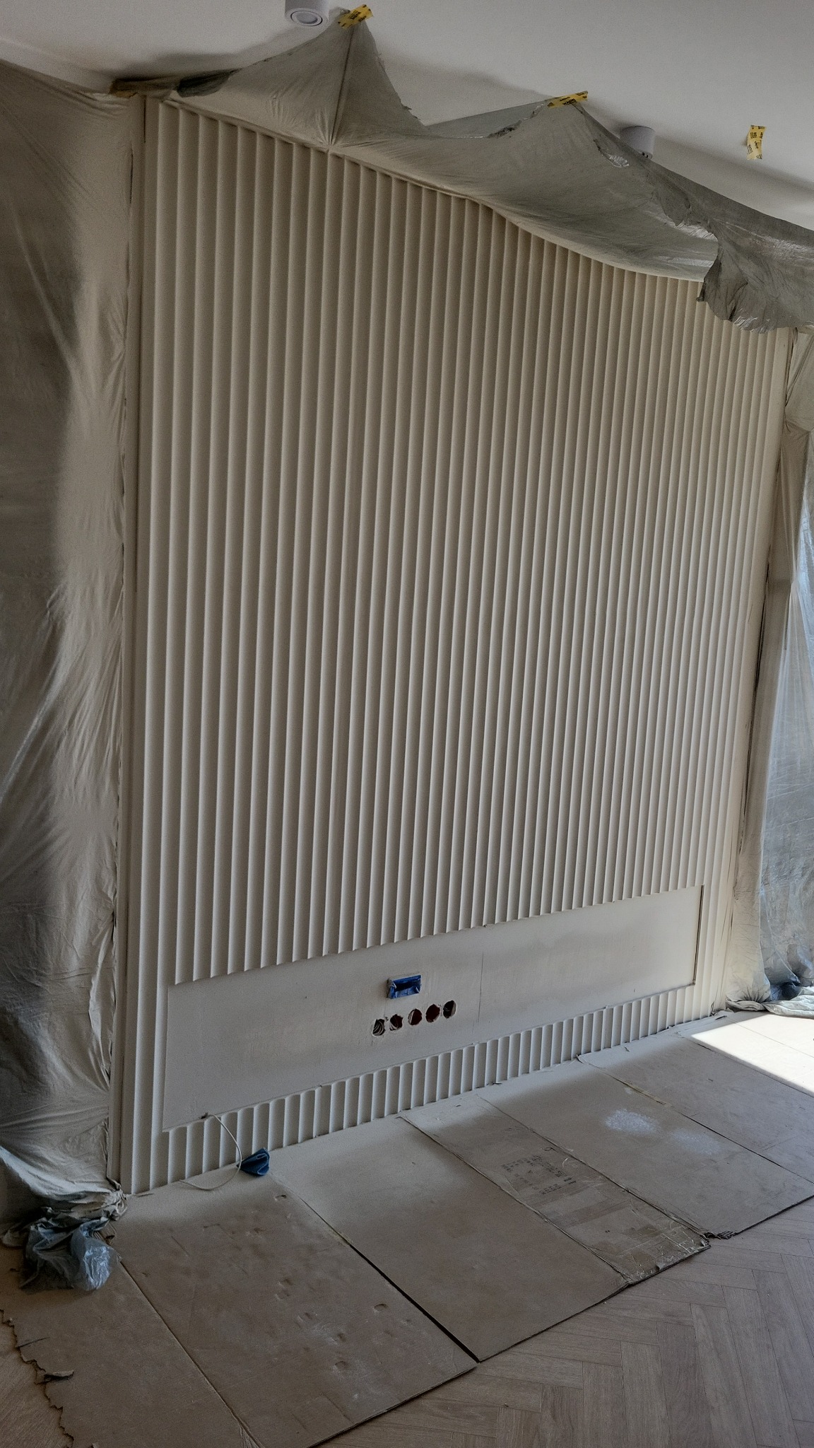 Ściana z dekoracyjnymi, pionowymi panelami w trakcie montażu, zabezpieczona folią malarską, widoczne gniazdka elektryczne i parkiet na podłodze.