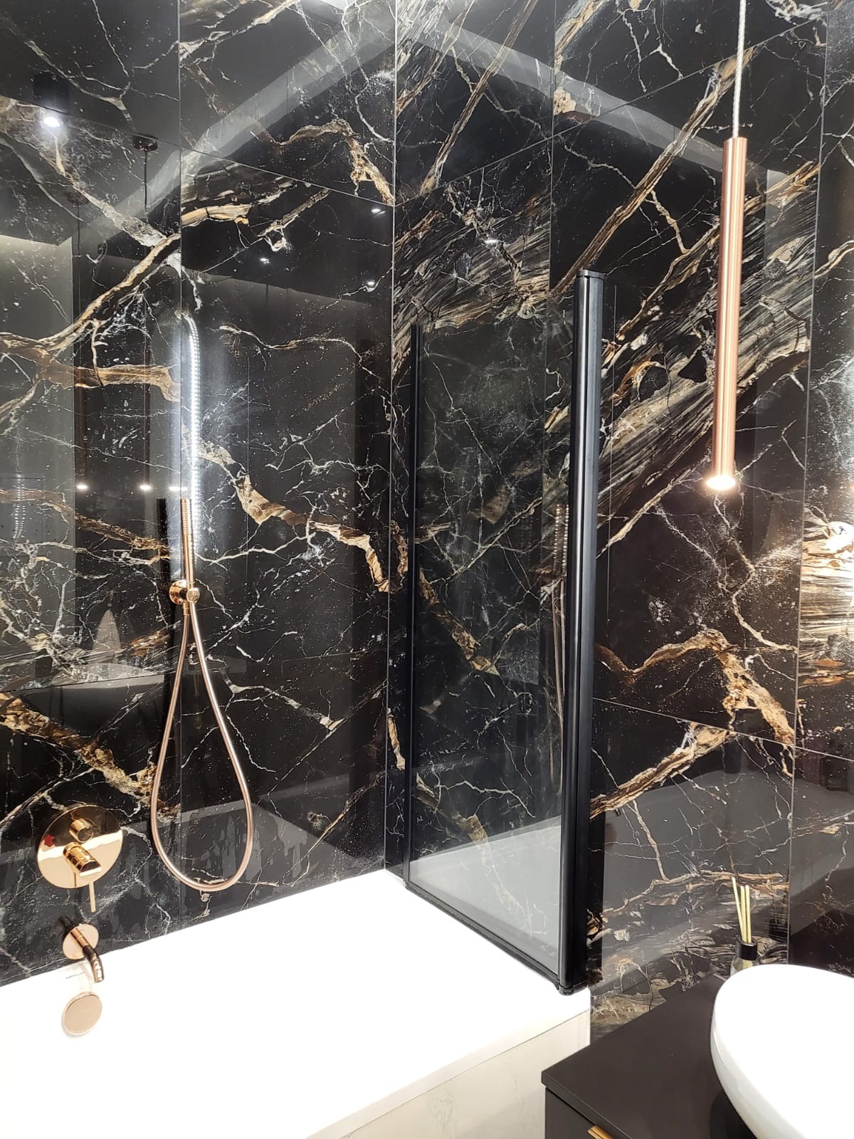 Luksusowa łazienka z czarnymi płytkami imitującymi marmur ze złotymi żyłami, armatura w kolorze różowego złota, szklana kabina prysznicowa z czarnymi profilami.