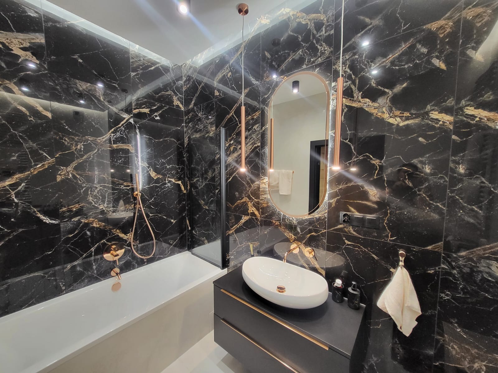 Elegancka łazienka z czarnymi płytkami imitującymi marmur, armaturą w kolorze różowego złota i owalnym lustrem.