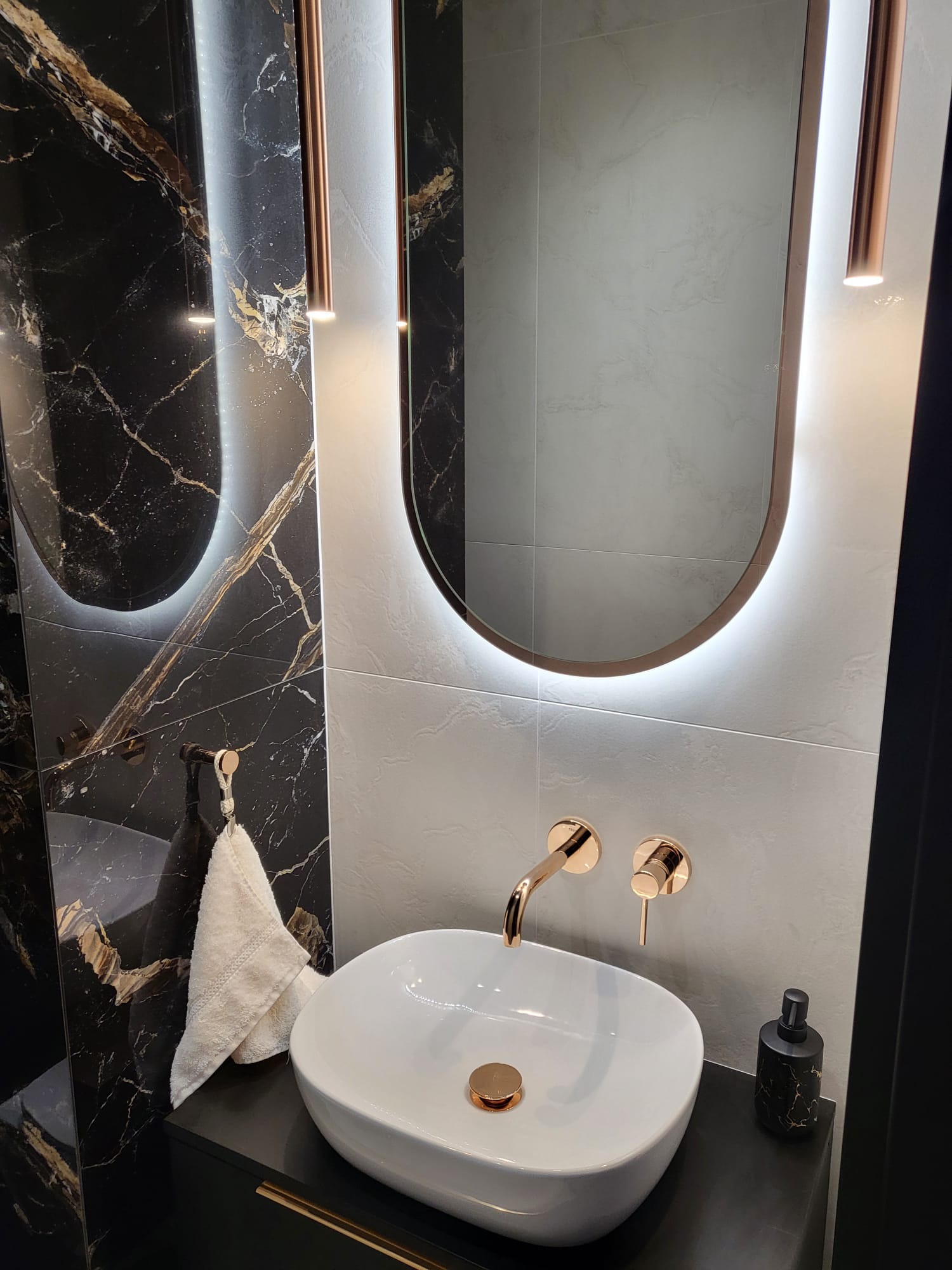 Nowoczesna łazienka z umywalką nablatową, złotą armaturą i podświetlanym lustrem na tle czarnych i szarych płytek imitujących marmur.