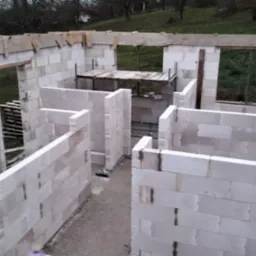 Wznoszenie ścian parterowego domu z bloczków betonu komórkowego; widoczne otwory okienne i drzwiowe, drewniane nadproża, rusztowanie wewnątrz budynku.