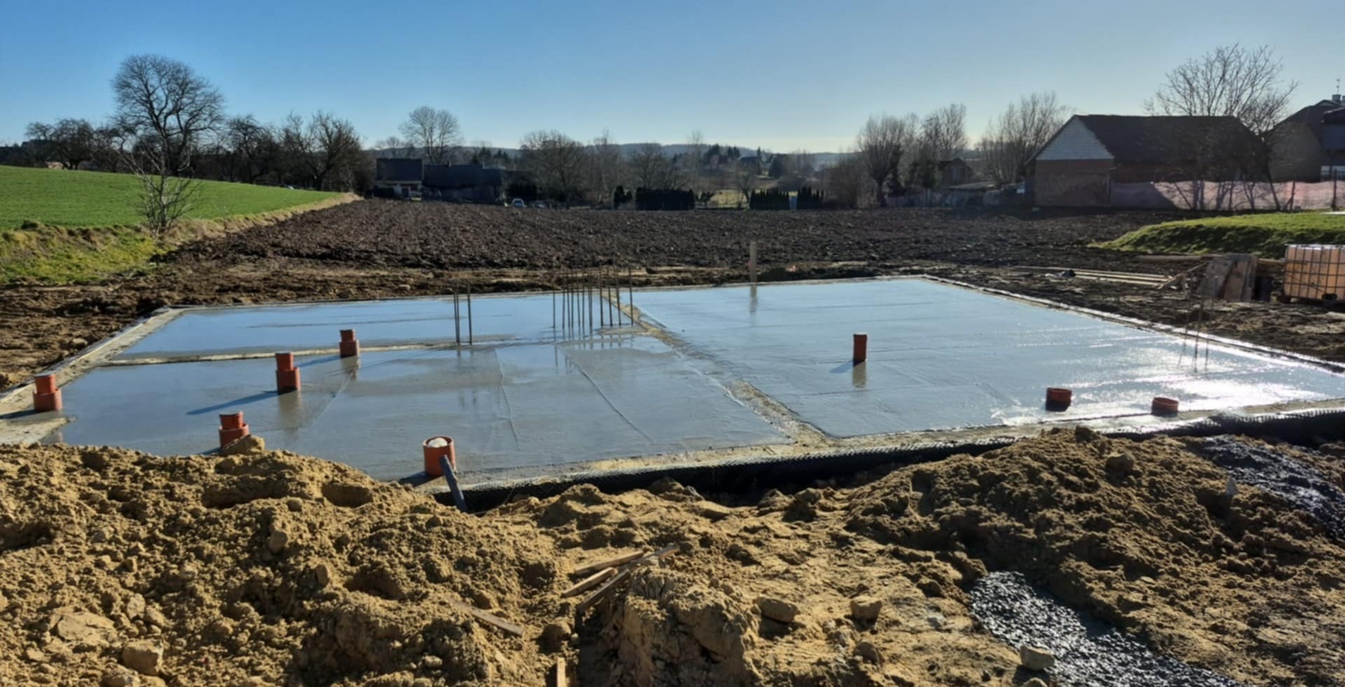 Wylany, mokry fundament płyty betonowej z wystającymi rurami kanalizacyjnymi na tle zaoranego pola i zabudowań wiejskich w słoneczny dzień.
