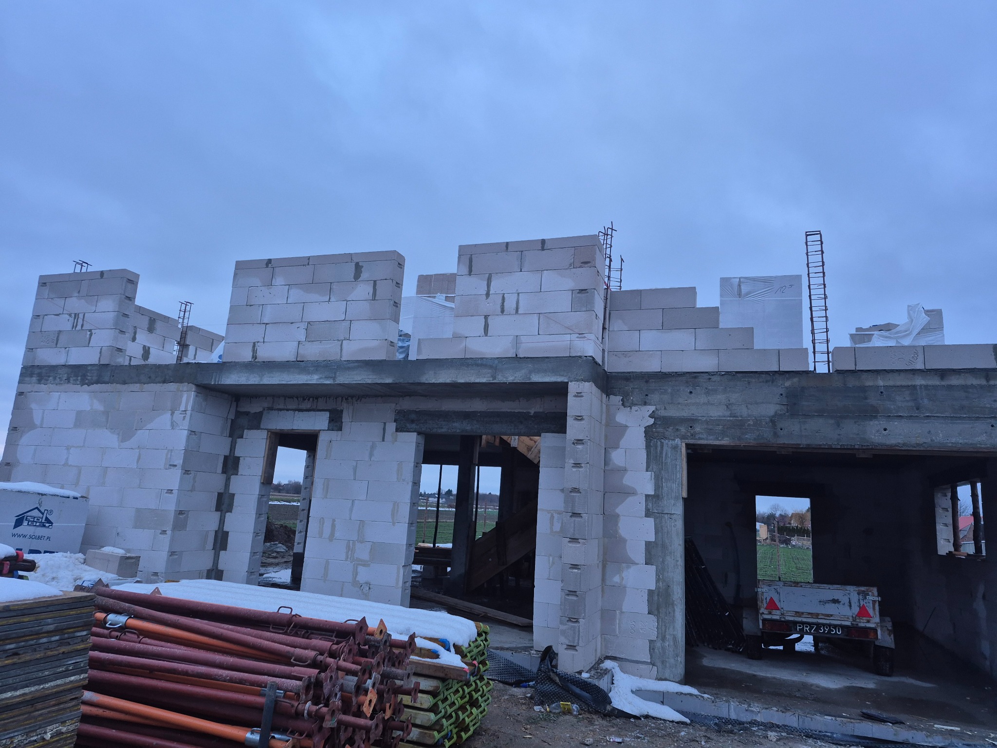 Wznoszenie ścian domu z białych bloczków betonu komórkowego. Widoczne zbrojenia i podpory. Na pierwszym planie stosy podpór budowlanych i przyczepka samochodowa.