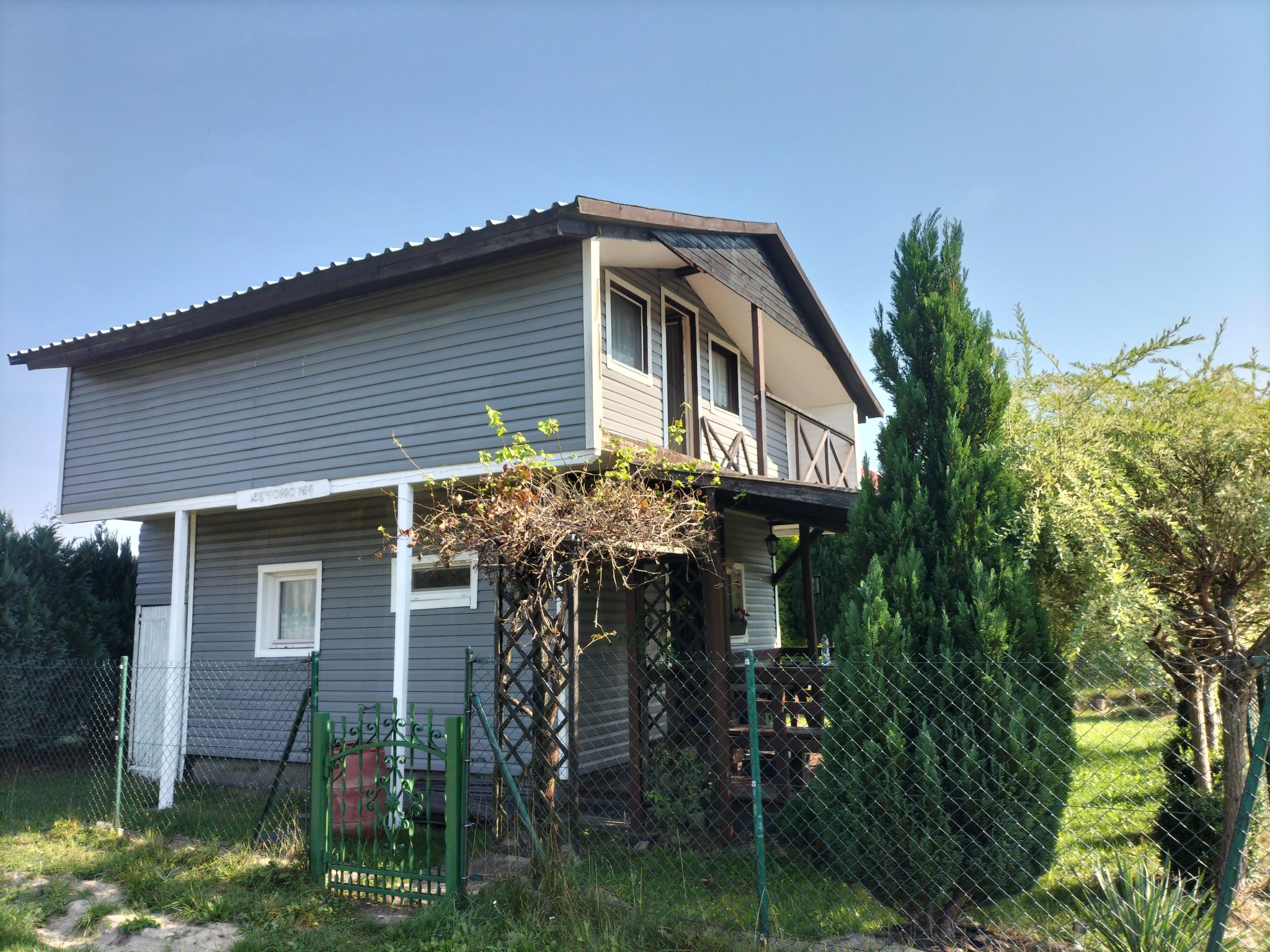 Dwukondygnacyjny dom z szarą elewacją, drewnianą werandą i ażurową konstrukcją wejściową porośniętą pnączami, otoczony zielonym ogrodzeniem z siatki.