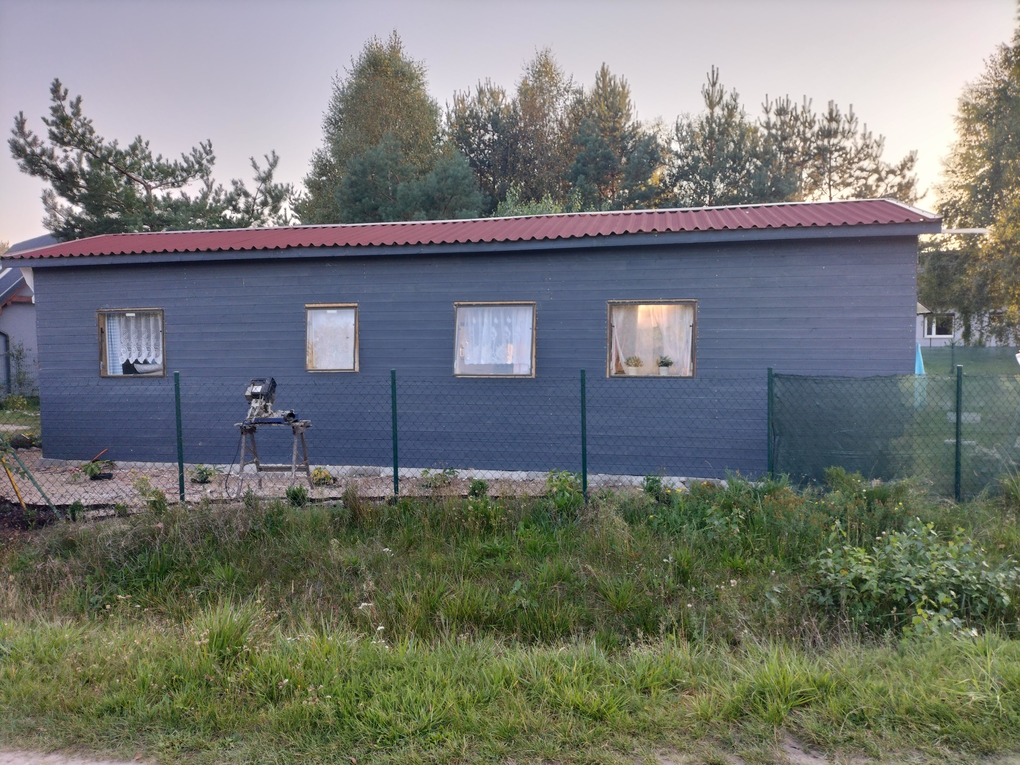 Szary budynek gospodarczy z czerwonym dachem, widoczny zza zielonego ogrodzenia, z trzema oknami z firankami i kwiatami w doniczkach na parapetach, przed budynkiem ustawiony statyw z narzędziami.
