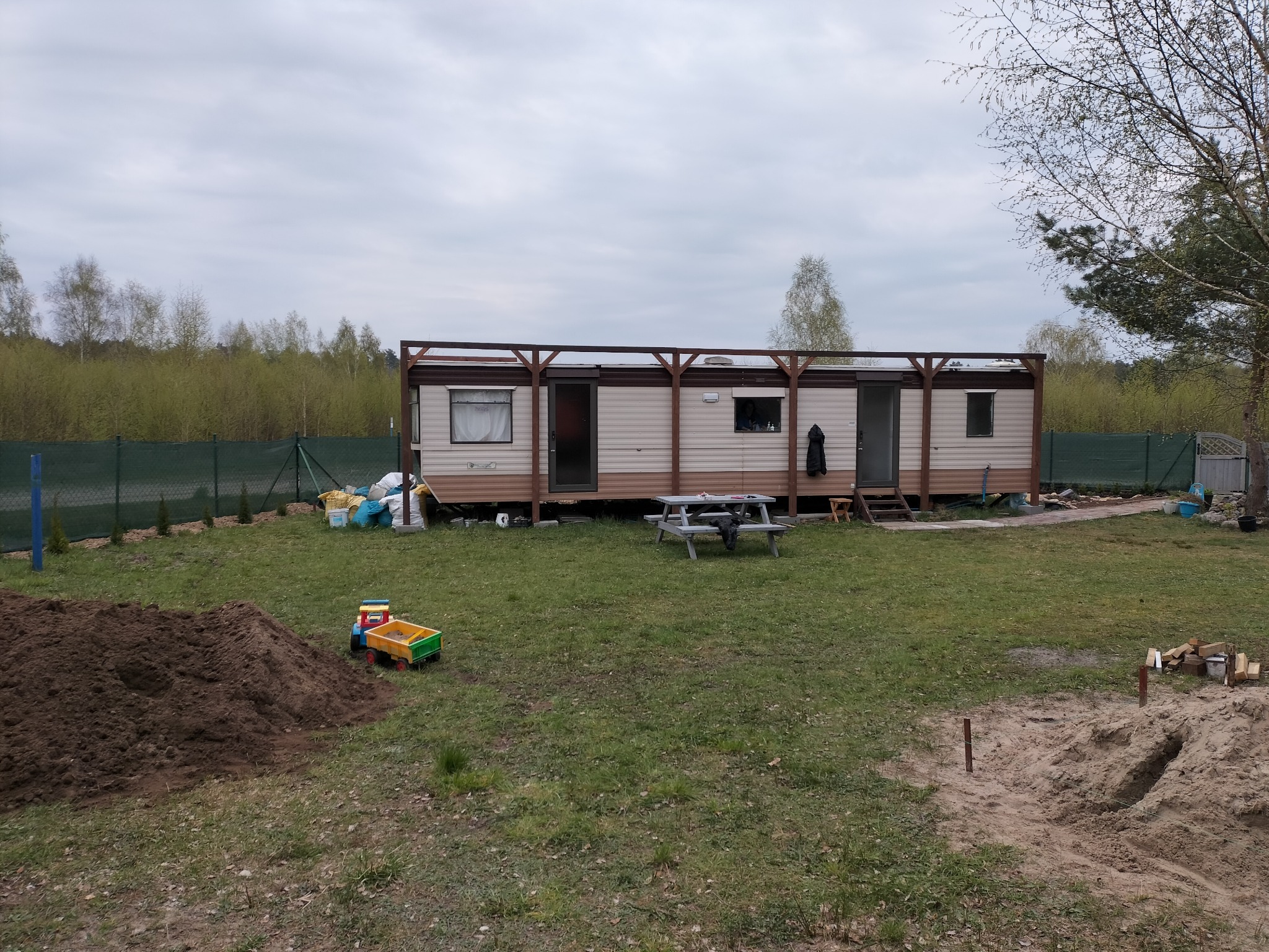 Mobilny domek z drewnianą konstrukcją zadaszenia, widoczny stół piknikowy, kupka ziemi, zabawki dziecięce i przygotowane drewno na opał na działce z ogrodzeniem z siatki.